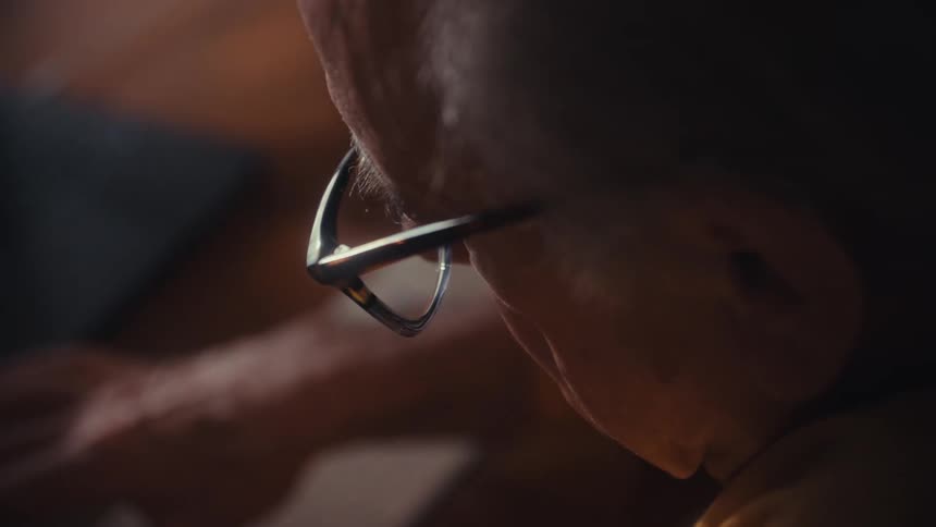 An over-the-shoulder close-up of a senior person with glasses looking down at their desk while working in a dimly lit room