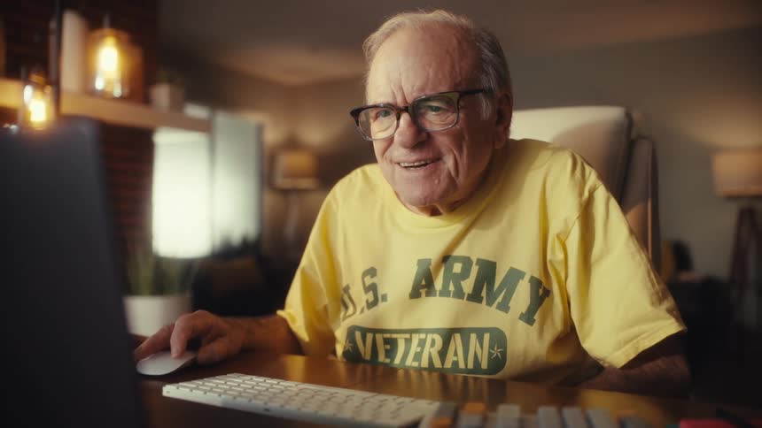A happy U.S. Army veteran wearing glasses smiles and laughs while working on his laptop in his cozy home office