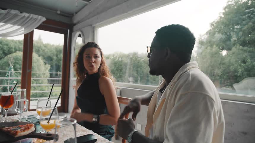 A diverse young couple chats animatedly while having a meal at a bright, modern restaurant with a view
