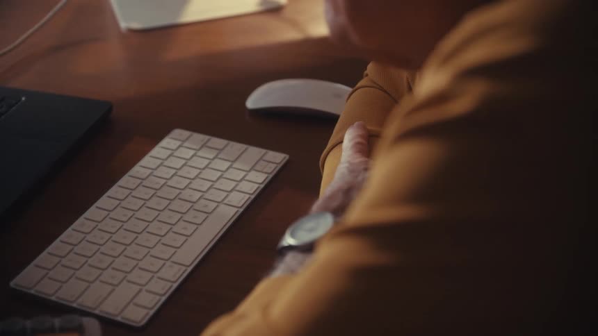 Close-up over the shoulder shot of a senior man with glasses typing on a computer keyboard while working at his desk