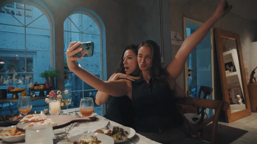Two joyful female friends take playful selfies with a smartphone while enjoying dinner at a stylish restaurant