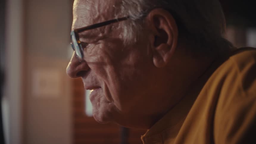 A close-up side profile of a smiling elderly man with glasses enjoying content on a screen in a warmly lit room