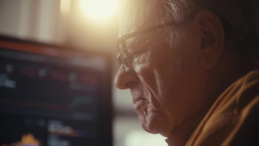 Close-up of a focused elderly man in glasses analyzing stock market charts on a computer screen with warm backlighting