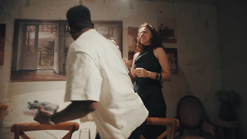A man is ecstatically surprised by a gift from his two female friends during a dinner party, leading to a joyful group hug