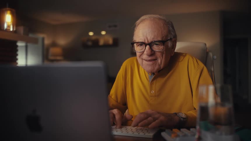 A senior man with glasses sitting at his home office desk experiences a moment of joy and surprise while using his computer