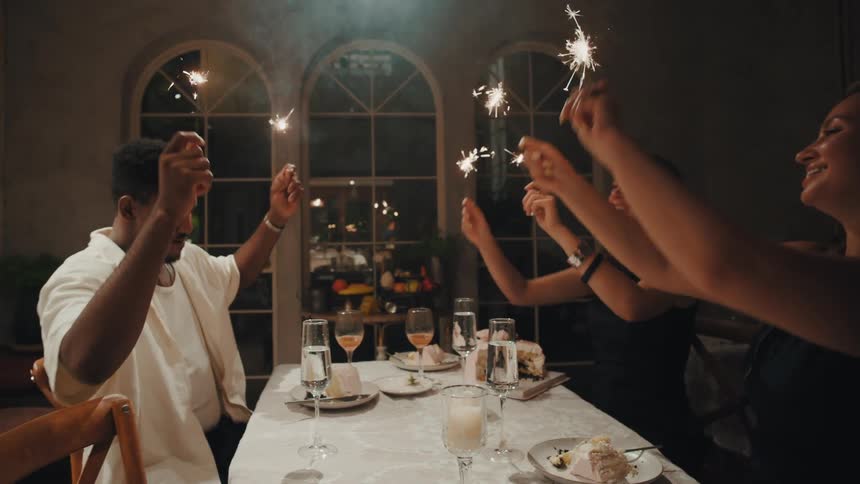 A group of happy friends celebrate a special occasion by laughing and waving sparklers at a dinner party in slow motion
