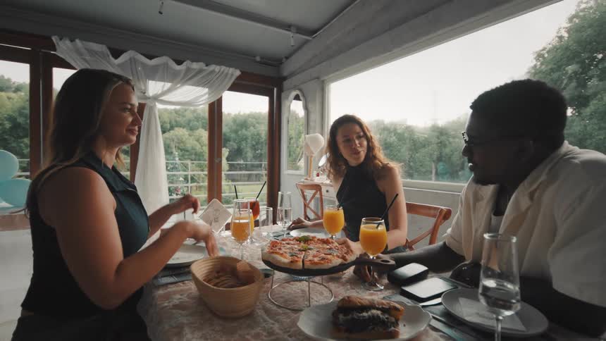 Happy multiethnic friends toast with orange juice while dining and laughing together in a beautiful restaurant with a view