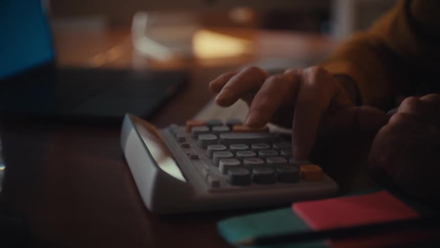 An elderly man in glasses sits at his desk in a dimly lit room, intently using a calculator to manage his finances