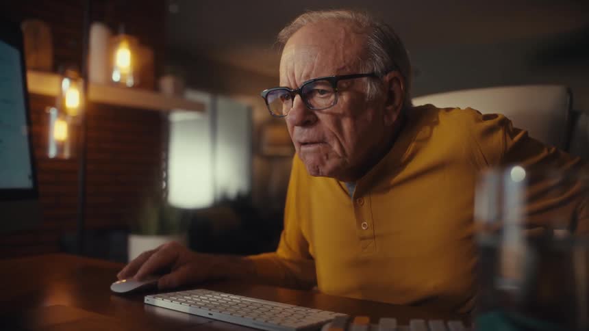 An elderly man with glasses works on his computer in his home office at night, concentrating on the screen before breaking into a smile
