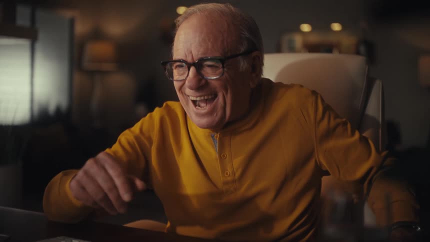 Elderly man with glasses enjoys a heartwarming and funny video call on his laptop, laughing joyfully in his dimly lit living room at night