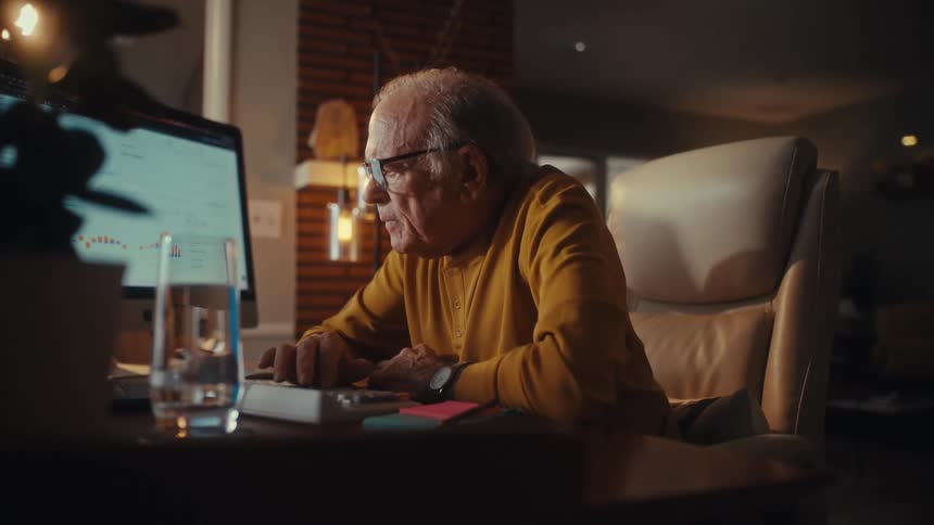 A serious elderly man wearing glasses manages his finances late at night in his home office, using a calculator and checking data on his computer screen