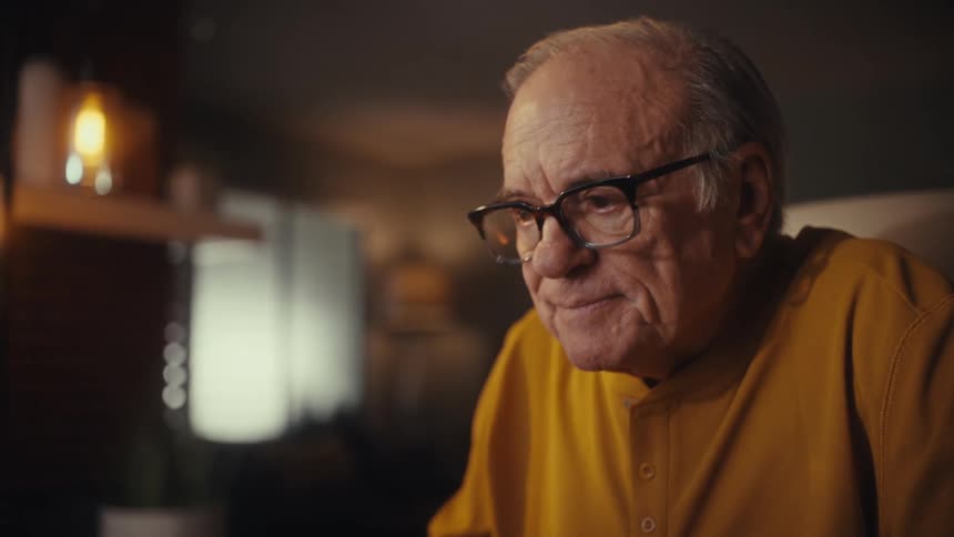 A senior man with glasses smiles while watching a screen, then focuses intently as he learns to type on his computer keyboard in a warmly lit room at night