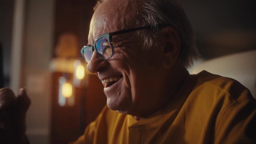 A delighted senior man in glasses celebrates a successful financial outcome with a joyful laugh while working late at his home office desk