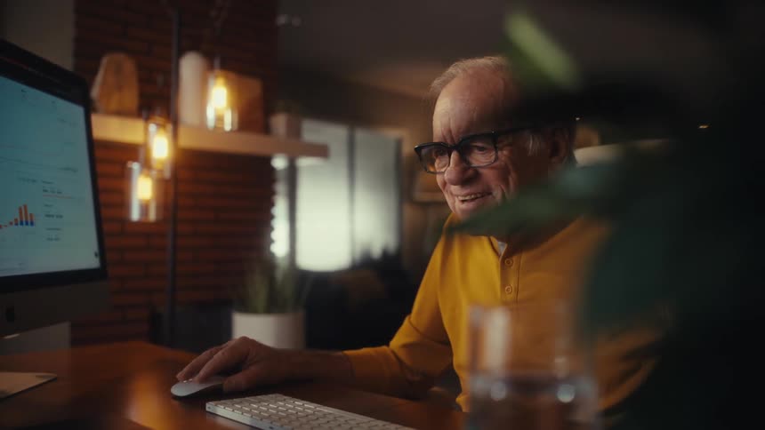 Happy senior man in glasses learns to navigate a computer at his desk in a cozy home office at night