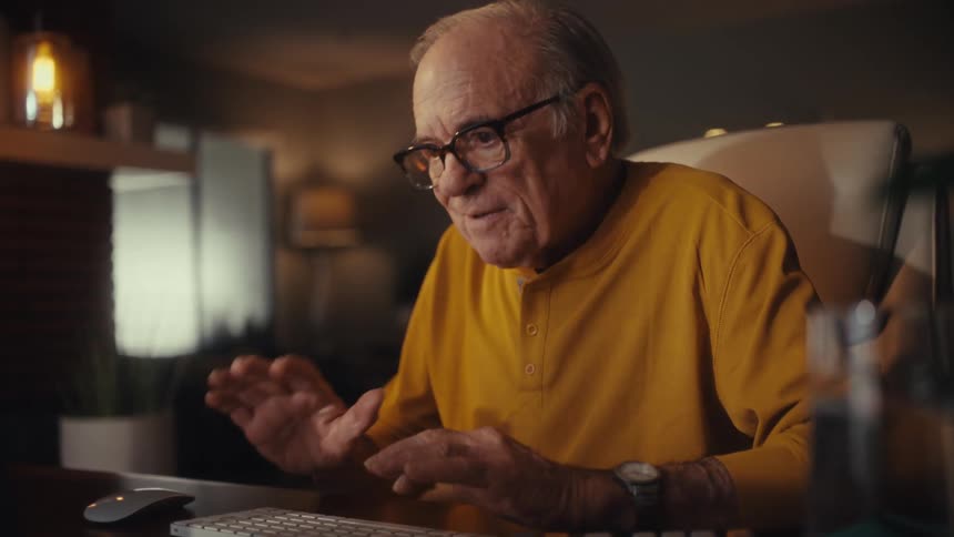 A senior man with glasses carefully types on his computer keyboard at night in his cozy home office and smiles with satisfaction