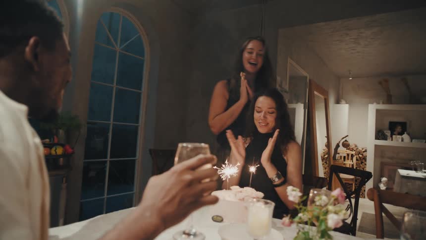 Joyful woman brings a birthday cake with sparklers to surprise her friend at a lively dinner party