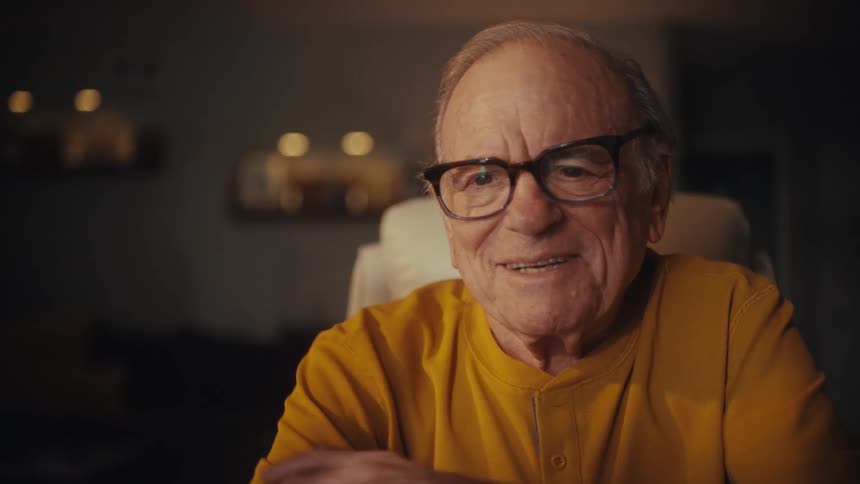 A charming senior man in a yellow shirt laughs and waves while on a video call, happily taking off his glasses at his desk