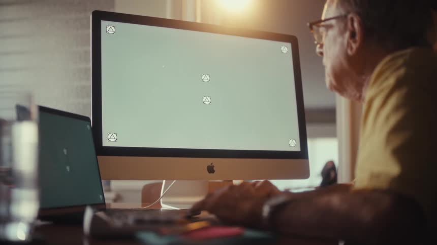 An elderly man with glasses works from home, typing on his computer with a green screen monitor