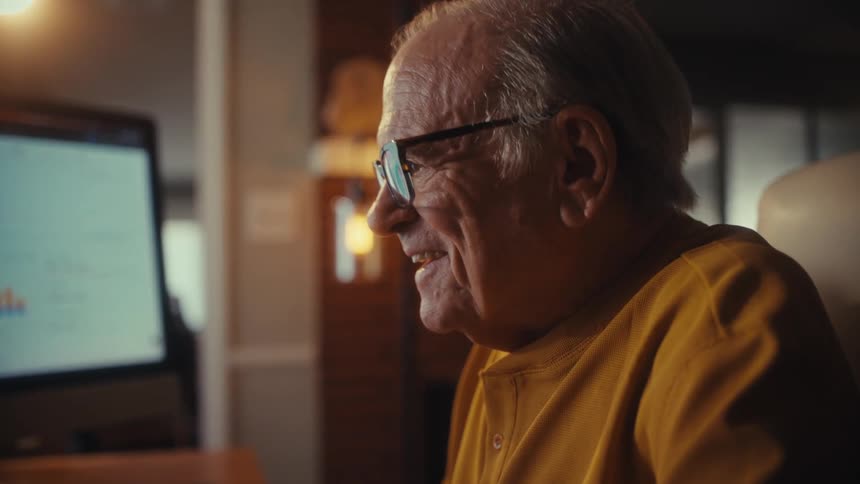 A smiling senior man with glasses works on his home computer, looking at charts with a look of satisfaction