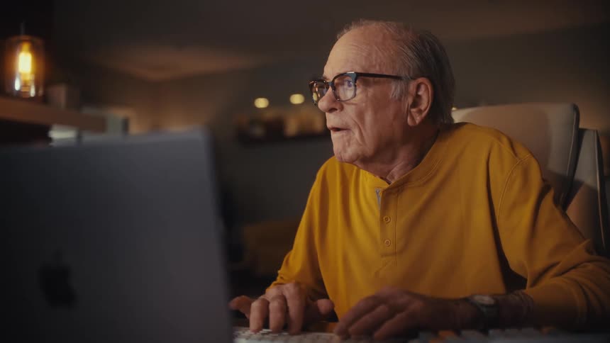 Joyful elderly man with glasses smiles while typing on his laptop in a cozy, warmly lit room at night