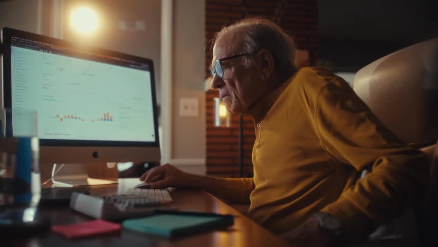 Focused senior man in glasses reviews business data and graphs on his computer in a warmly lit home office