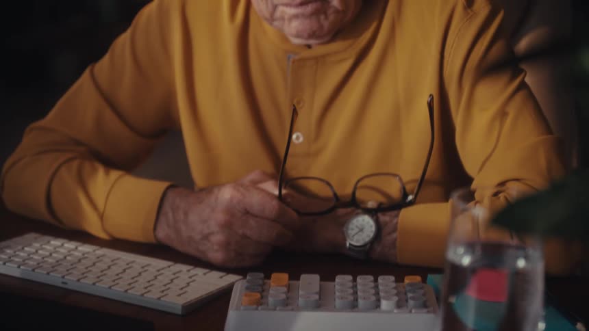 A senior man in a yellow shirt works on his computer at night, pausing to take off his glasses with a pensive look