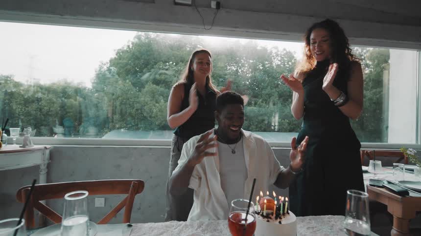Joyful friends surprise a young man with a birthday cake in a sunlit restaurant, leading to a happy celebration of him blowing out the candles