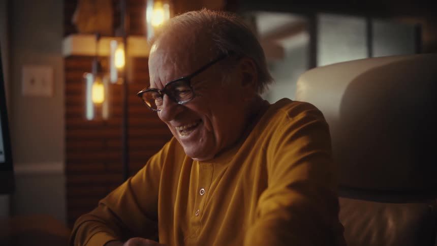An overjoyed elderly man laughs uncontrollably and dances in his chair while watching something on his computer at home