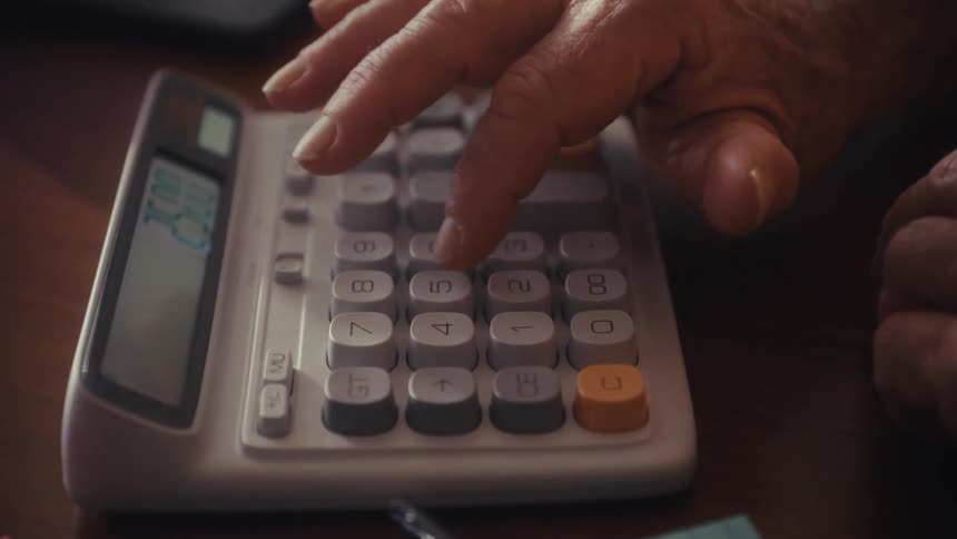 Close-up of an older person's hands using a calculator and typing on a keyboard at a desk