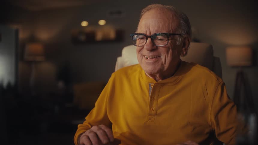An elderly man in glasses and a yellow shirt smiles and laughs while learning to use his computer at night in his cozy home office