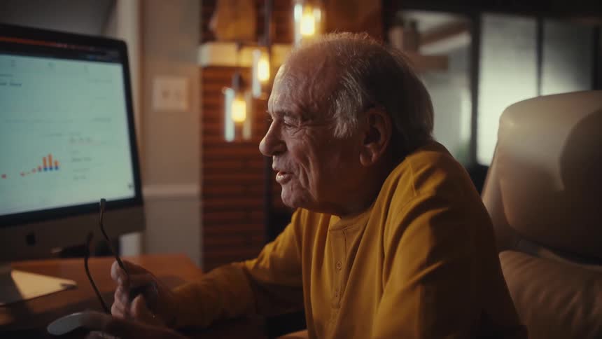 A happy elderly man celebrates financial success while on a video conference call in his home office at night
