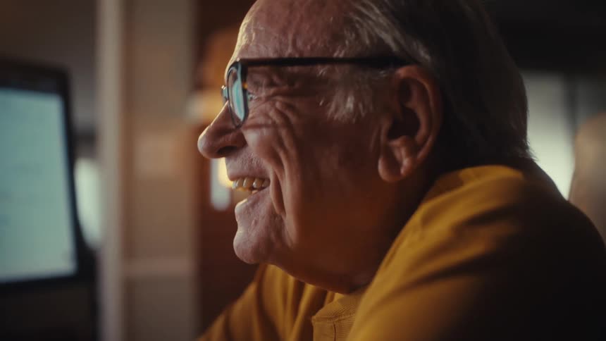 An elderly man in a yellow shirt smiles and celebrates a success while working on his computer in a cozy home office at night