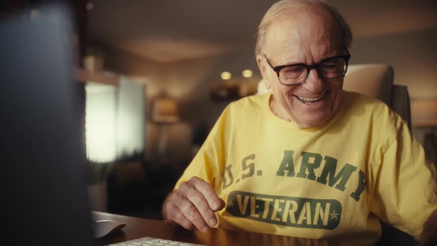 A happy elderly U.S. Army veteran smiles and waves while on a video call on his computer, then uses a calculator at his desk in his home