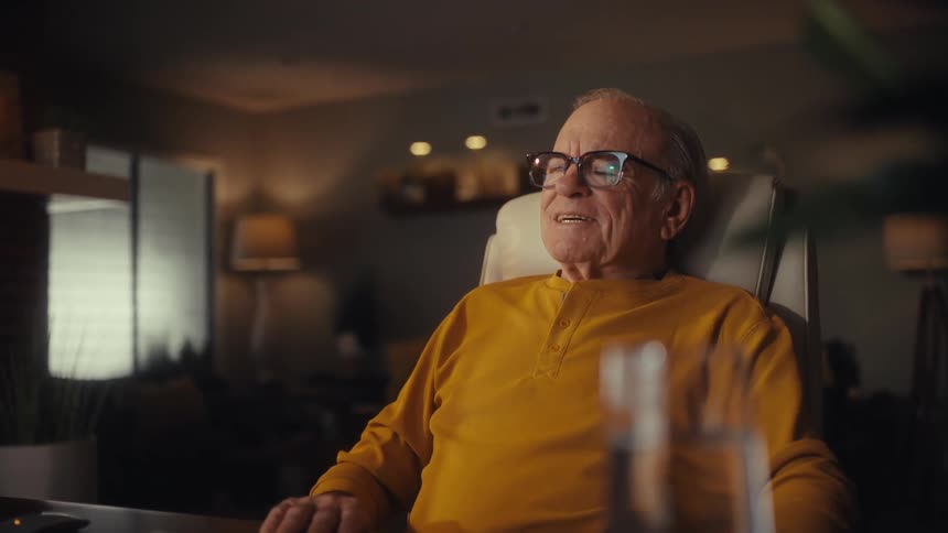 An elderly man in glasses and a yellow shirt smiles with relief after finishing his work on the laptop in his home office at night