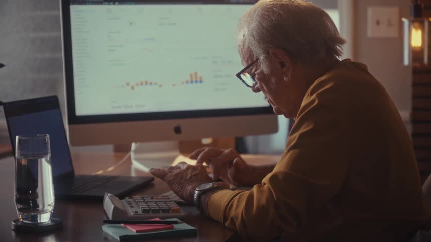 A senior man with glasses reviews his investments on his computer, using a calculator, and happily celebrates a successful outcome in his home office