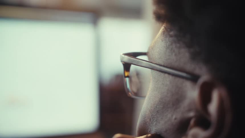 Over-the-shoulder shot transitions focus from a man wearing glasses to a computer screen displaying financial data and charts