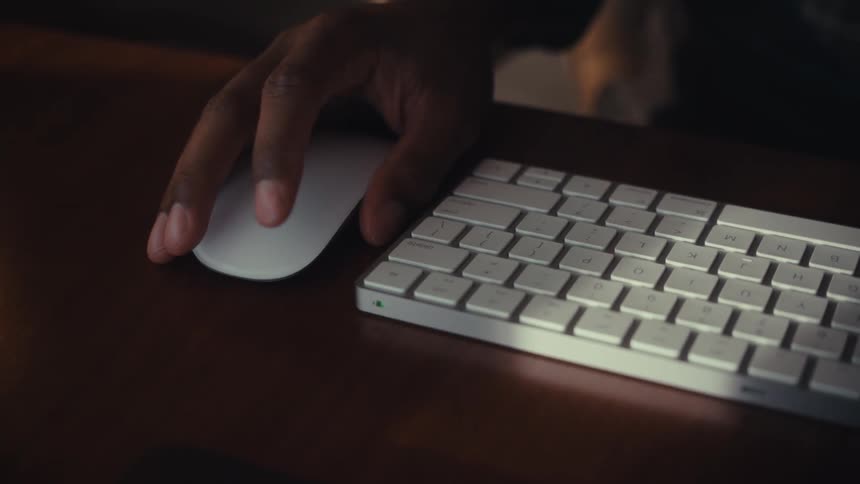 A dimly lit close-up of a hand using a white wireless mouse on a wooden desk, panning to a keyboard