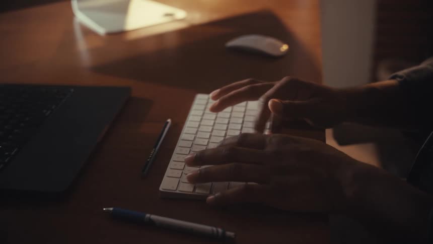 A slow pull-out shot focuses on a person's hands typing on a wireless keyboard at a desk illuminated by a strip of sunlight