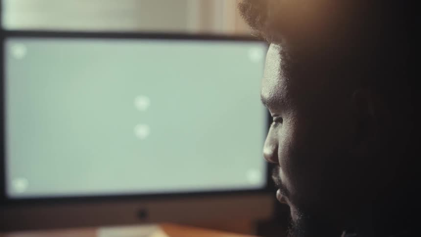 An over-the-shoulder slow push-in shot reveals an African American man intently focused on his desktop computer featuring a green screen