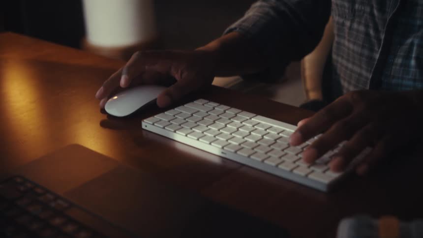 In a moody, dimly lit office, a man's hands move in slow motion across a keyboard and mouse as he works late into the night