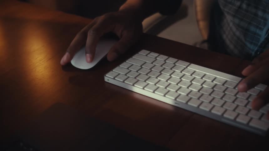 Close-up of a man's hands on a computer mouse and keyboard while working at a wooden desk in a dimly lit office