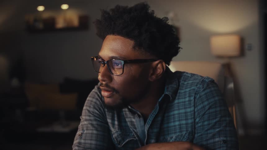 A young African American man with glasses transitions from a thoughtful expression to a smile while watching a screen at night