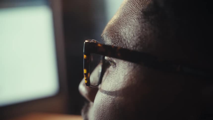 An extreme close-up of a Black man's eye reflecting a bright screen as he works intently in a dark room