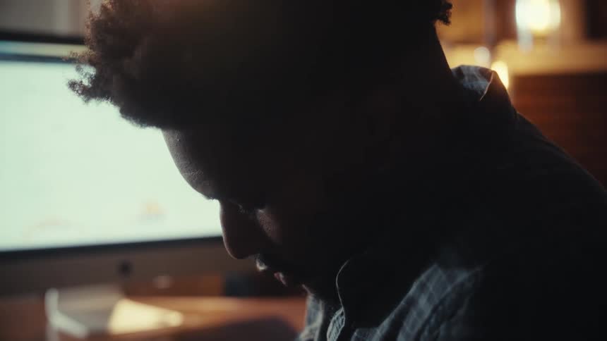 A focused man is silhouetted by his computer monitor as he writes notes at his desk while working late at night
