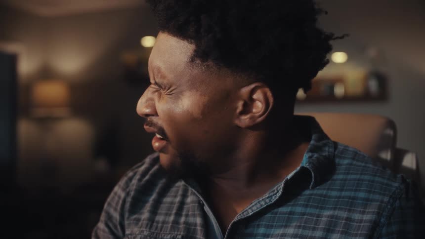 Slow-motion close-up of a tired young Black man in a plaid shirt yawning in a dimly lit room at night
