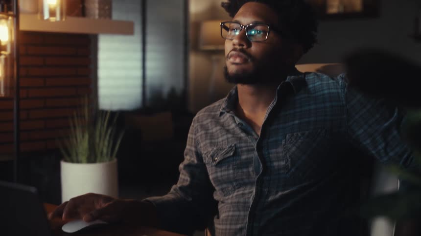 A focused young African American man wearing glasses analyzes data on his computer while working late from his cozy home office