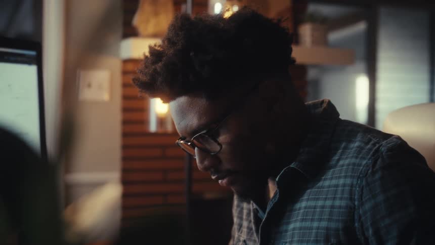 A focused African American man wearing glasses works on his computer at night in his cozy home office