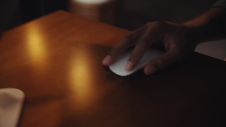 A close-up captures a person's hands as they type on a wireless keyboard and navigate with a mouse on a wooden desk under dramatic lighting