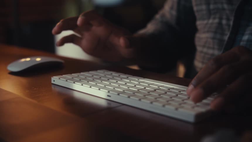 Close-up of a person's hands typing on a wireless keyboard and using a mouse at a wooden desk in a dimly lit room