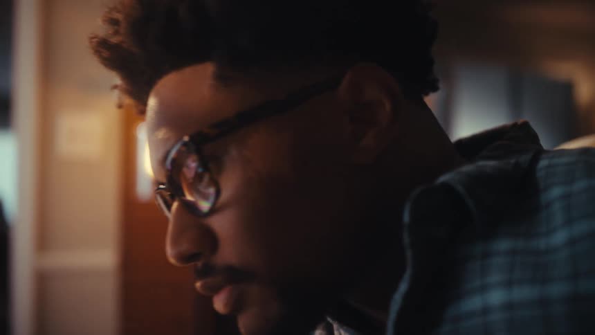 A pensive young man slowly puts on his glasses to continue working, with the screen reflecting in the lenses in a dimly lit room